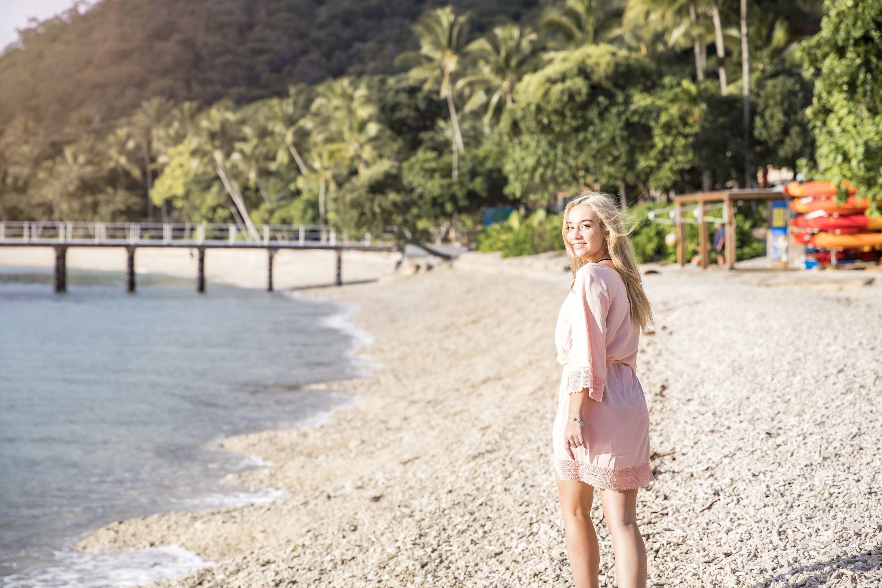 A beach scene with a wooden pier extending into the water, palm trees, and a person in a pink dress standing on the sand.