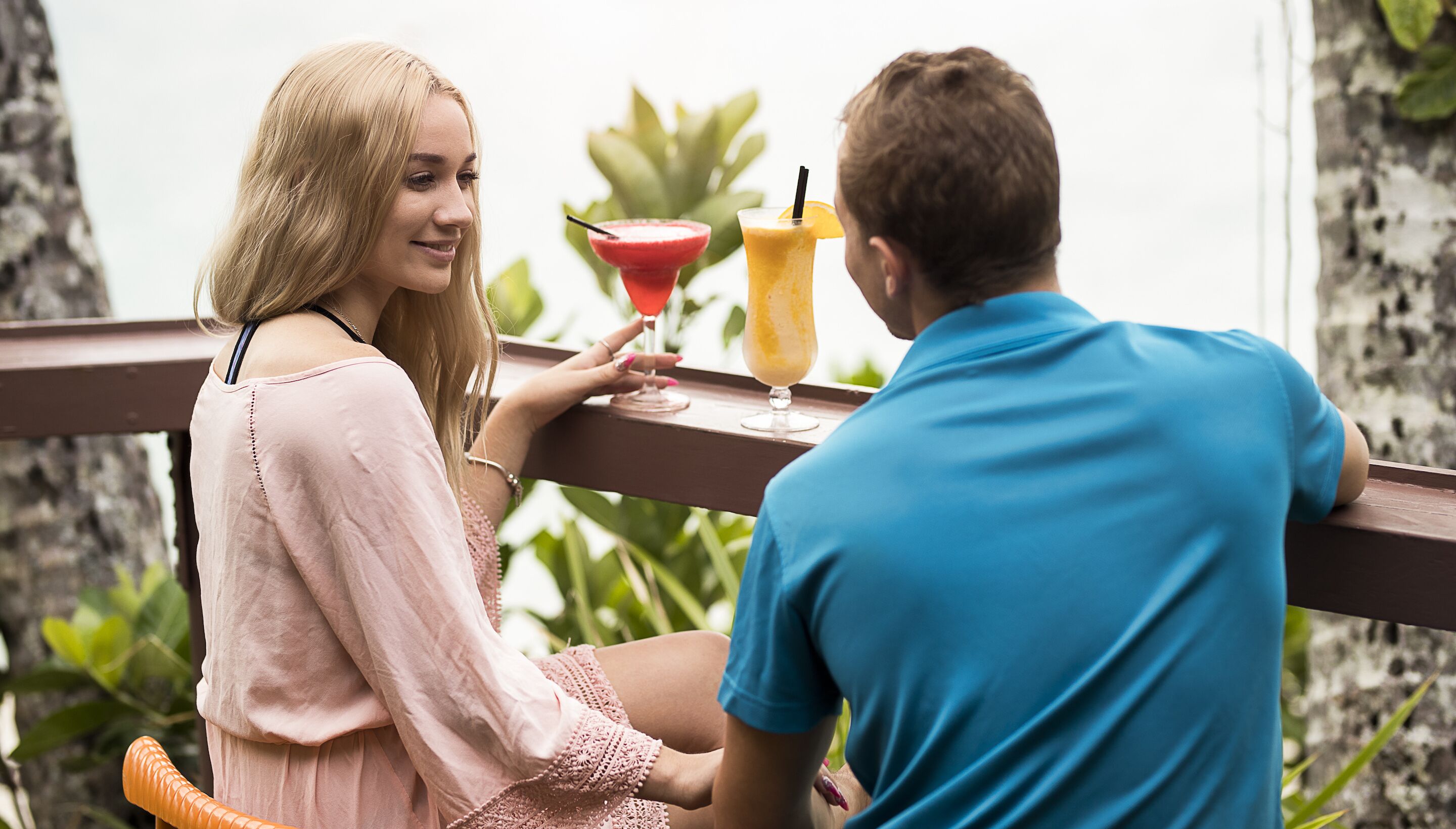 Two people seated at a wooden table with drinks, overlooking a garden with trees and a white wall.