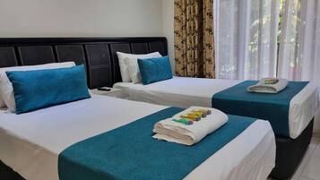 Habitación de hotel con dos camas, cada una con cubrecama color turquesa y ropa de cama blanca. Hay dos almohadas en cada cama, una turquesa y una blanca. Se ve un control remoto en la mesita de noche.