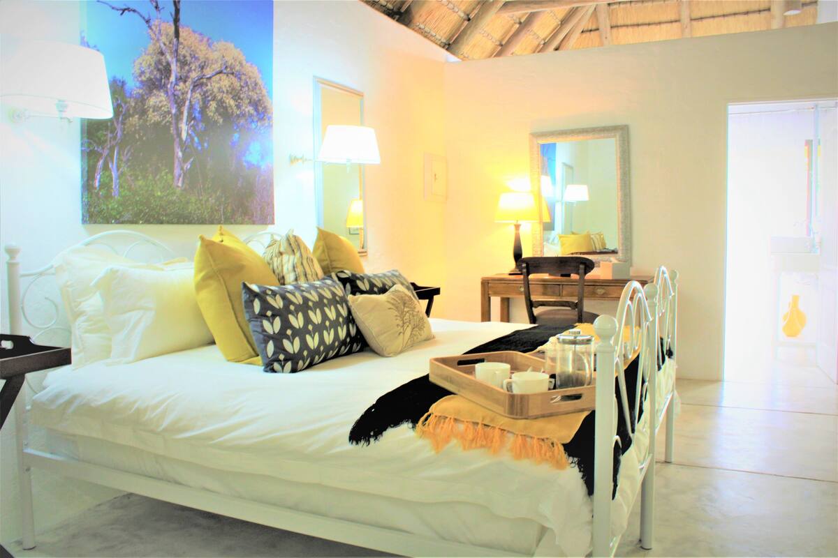 Un dormitorio con cama, almohadas, una mesita de noche, lámpara, espejo y ventana con vista a árboles.