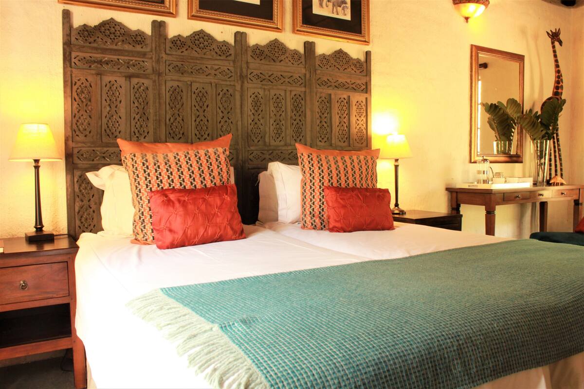 Una cama bien tendida con almohadillas estampadas, un cabecero decorativo, lámparas de noche, una mesita de noche y un espejo con adorno de jirafa.