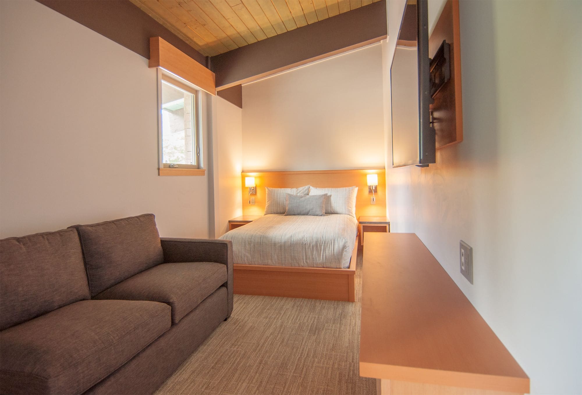 A bedroom with a bed, a wooden headboard, a nightstand with a lamp, a brown sofa, and a wooden bench.