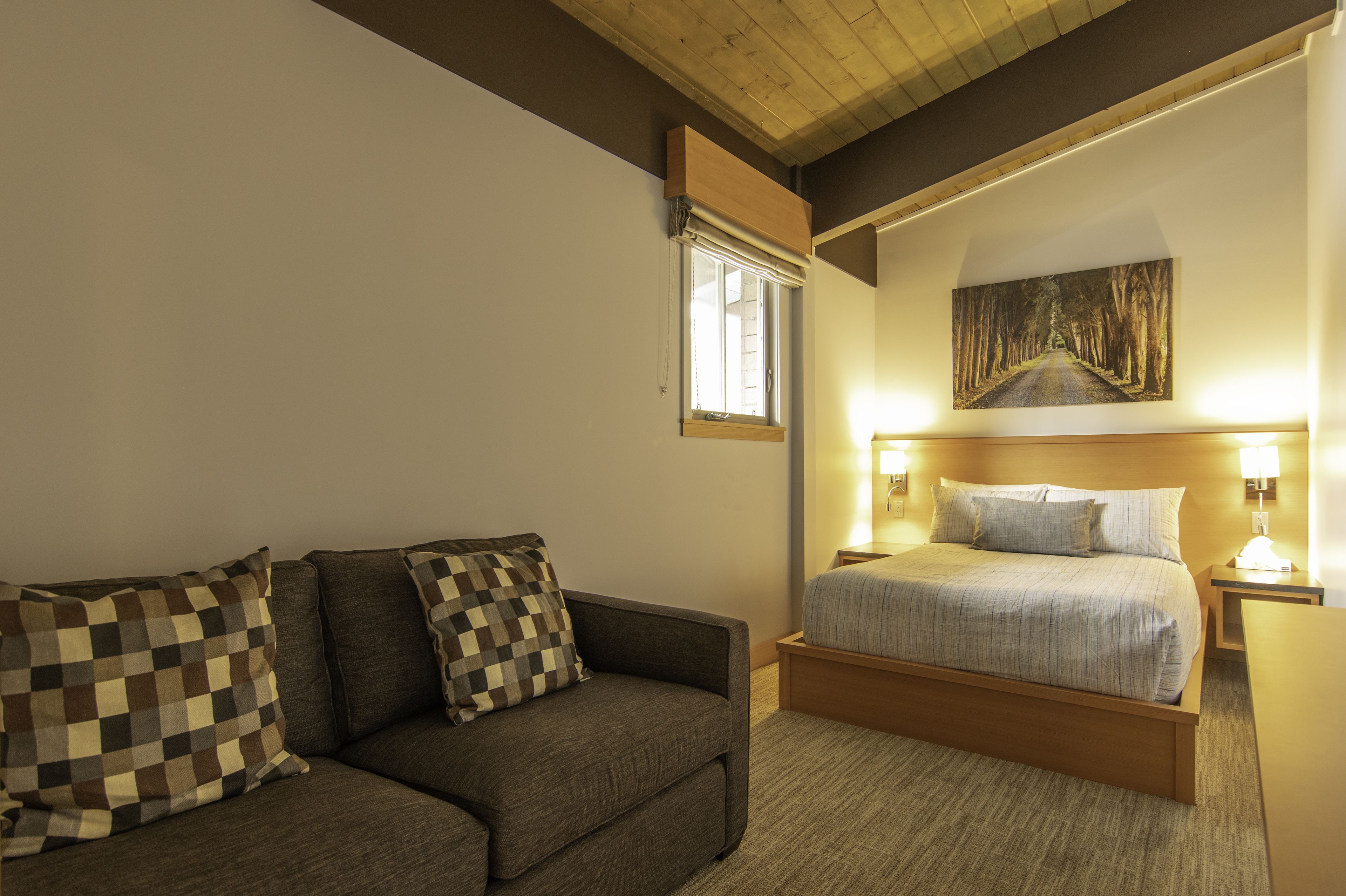A bedroom with a bed, a sofa, a window with blinds, a painting on the wall, and two bedside lamps.