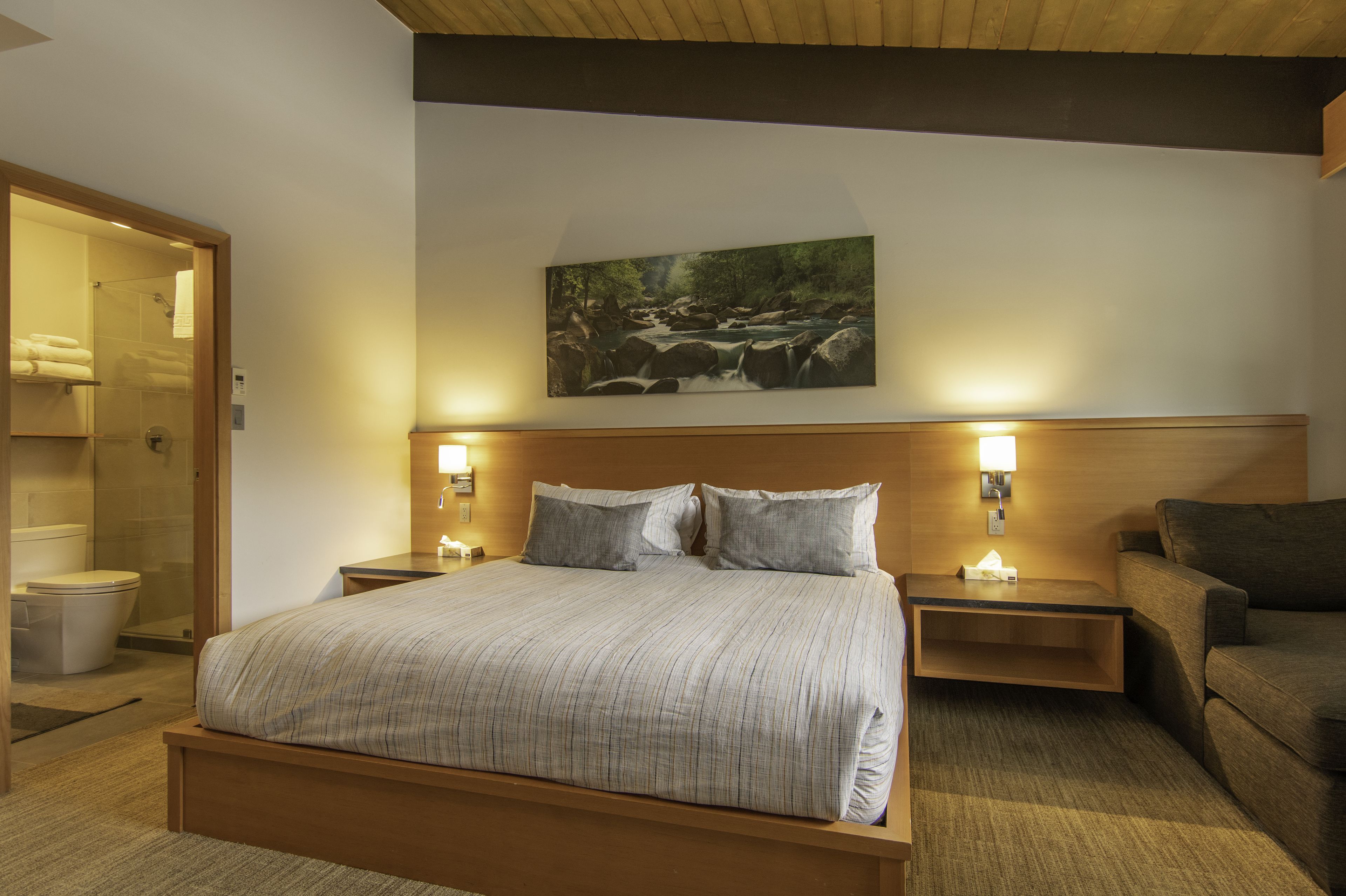 Ein Hotelzimmer mit einem großen Bett, zwei Nachttischen inklusive Lampen, einem Holzkopfschild, einem Gemälde einer Flusslandschaft und einer Holzbank.
