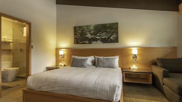 Une chambre d’hôtel avec un grand lit, deux tables de chevet avec des lampes, une tête de lit en bois, un tableau représentant un paysage fluvial et un banc en bois.