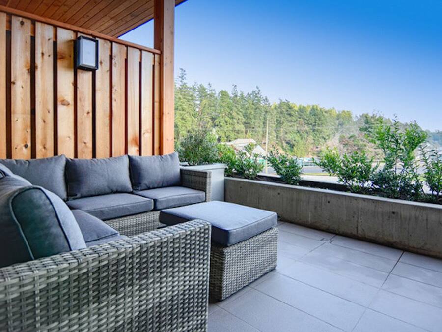 A covered outdoor seating area with a wicker sofa and ottoman, a wooden ceiling, and a view of a forested area.