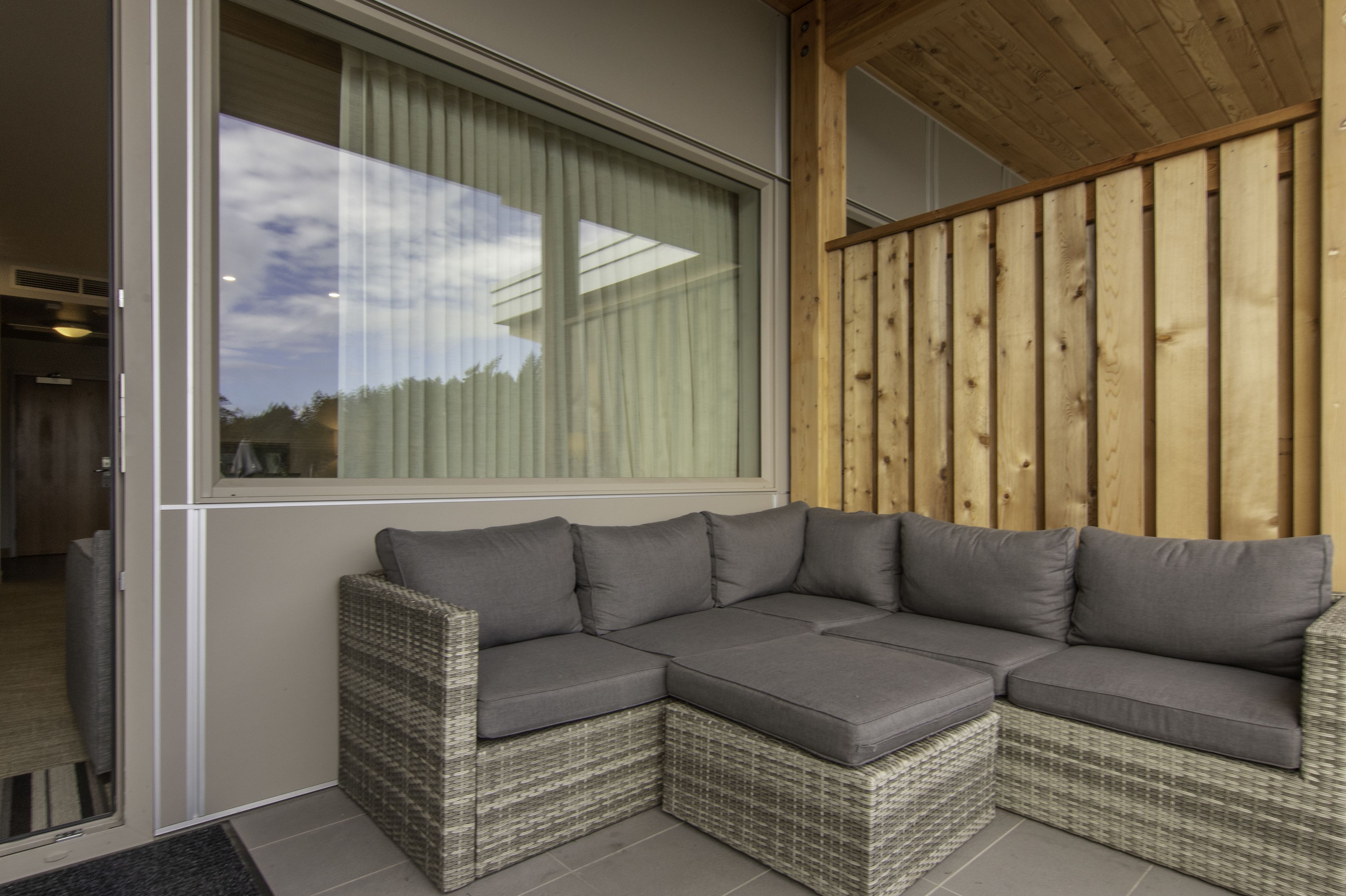 A corner sectional sofa with grey cushions, a wicker frame, and a view through a large window with curtains.
