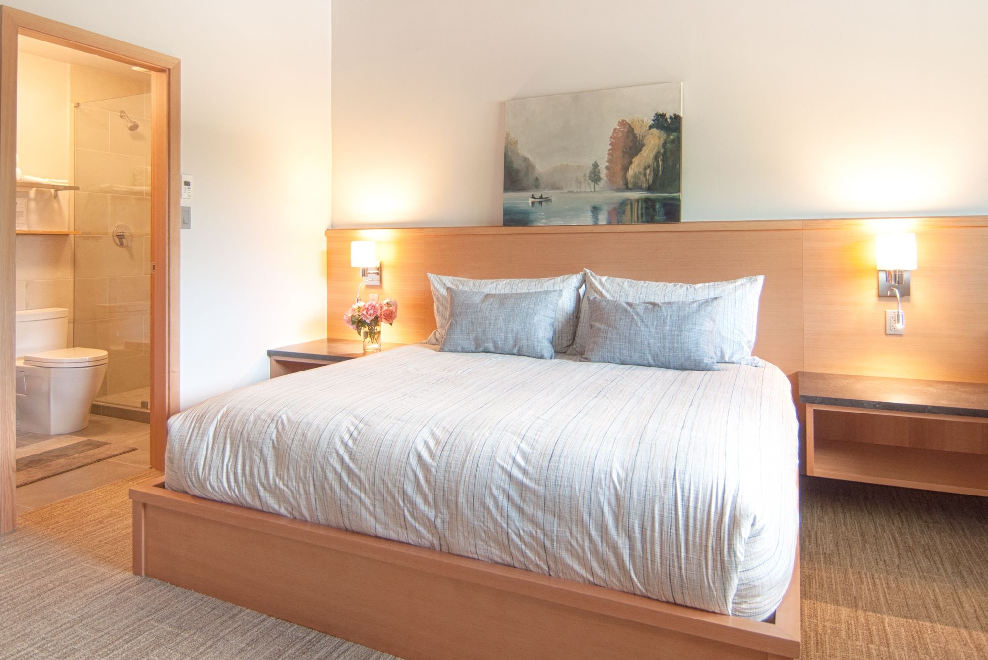 Habitación de hotel con una cama grande, cabecero de madera, dos mesitas de noche con lámparas, un cuadro sobre la cama y un baño con ducha e inodoro.