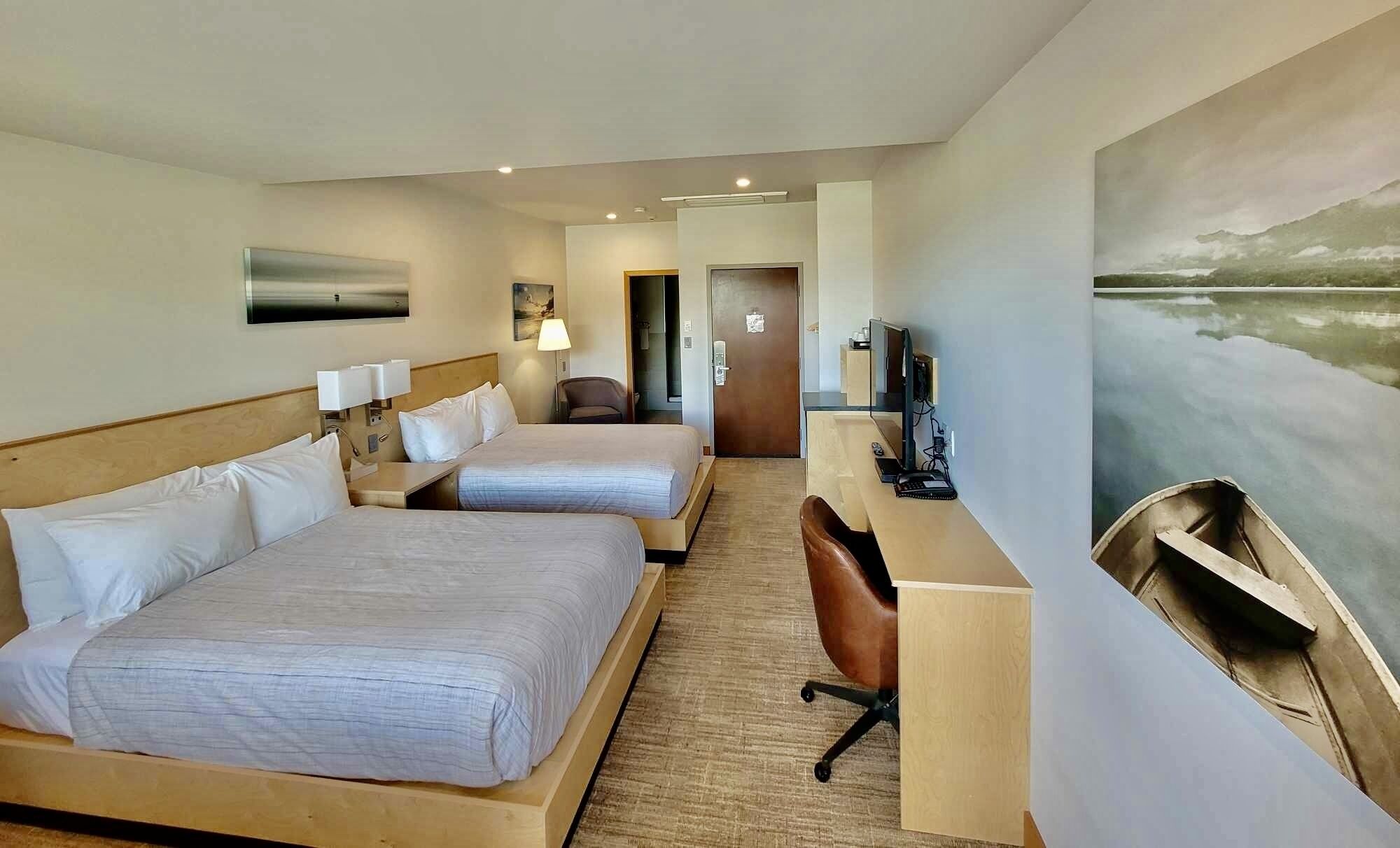 A hotel room with two beds, a desk with a computer, a chair, and a painting of a lake.