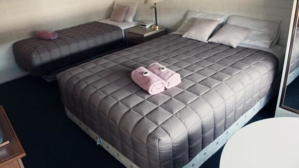 A hotel room with a large bed, two smaller beds, a bedside table with a lamp, and a round table. - H&D Apartments (St Kilda)