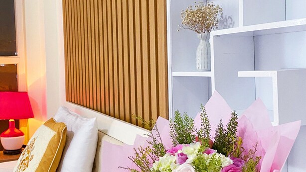 Een kamer met een wand van houten latten, een gitaar op een wit schap, een bloemenboeket en een bank met kussens.