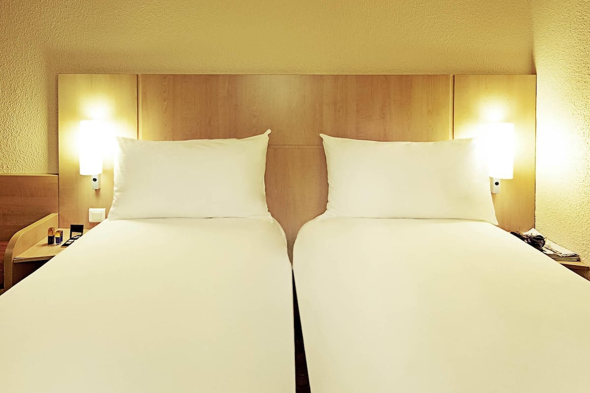 A hotel room with two beds, wooden headboard, bedside lamps, and a nightstand.