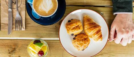 Et træbord med et fad croissant, en kop kaffe med latte-kunst og et glas frugtsaft.