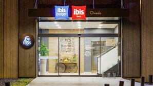 Entrada de hotel con señalización 'ibis budget' e 'ibis Oviedo', un aparcamiento para bicicletas y un letrero que dice 'Siéntete feliz en el bar'.