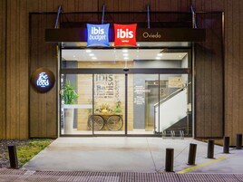 Entrada de hotel con señalización 'ibis budget' e 'ibis Oviedo', un aparcamiento para bicicletas y un letrero que dice 'Siéntete feliz en el bar'.