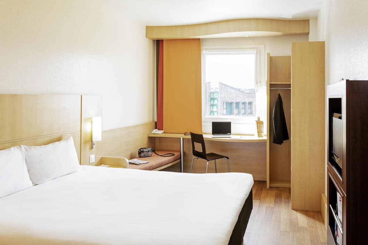 A hotel room with a large bed, a desk with a chair, a laptop, and a window with a view of buildings.