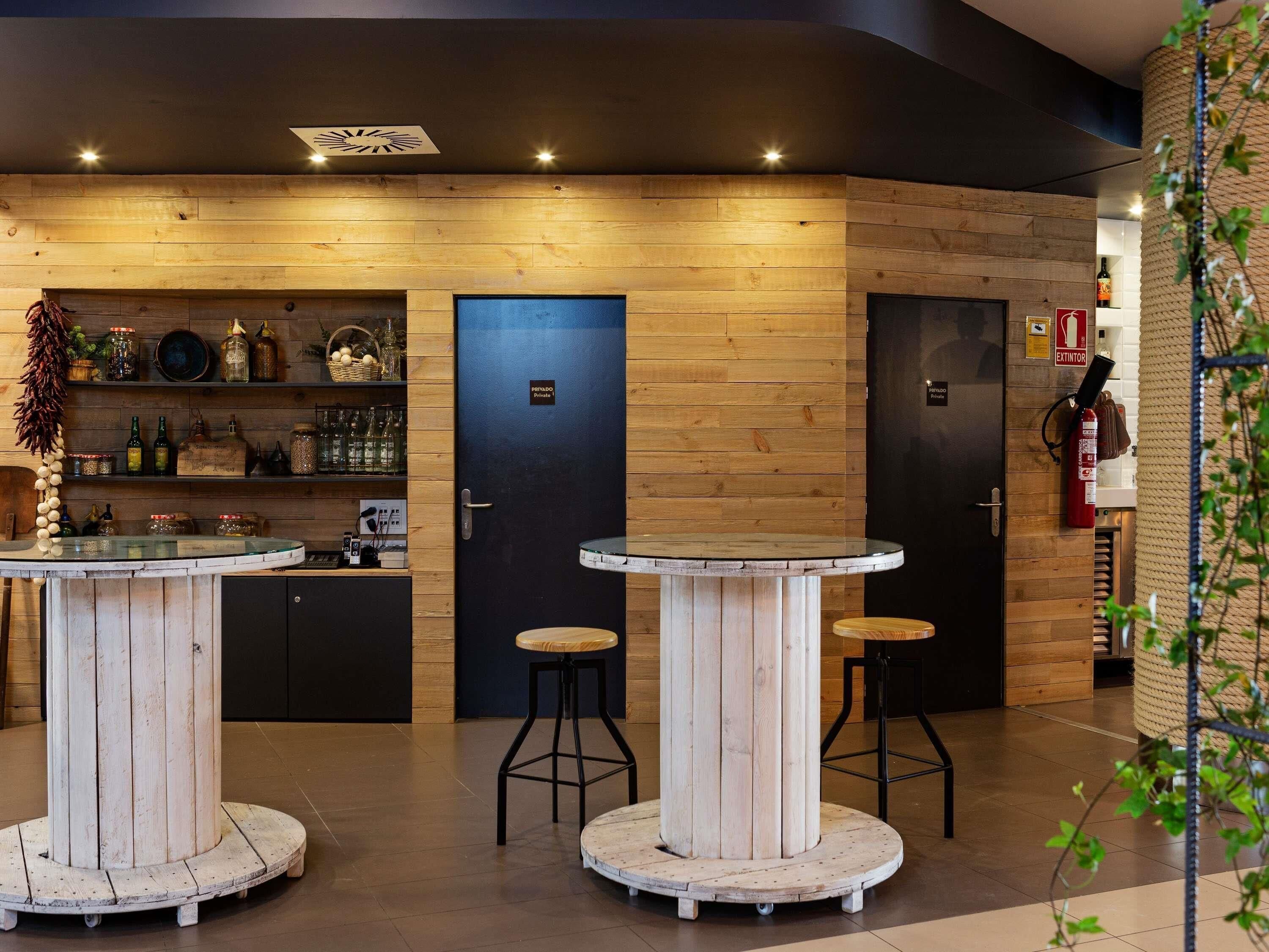 Un interior moderno con paneles de madera, dos mesas redondas con taburetes, una zona de bar con estantes llenos de botellas y un extintor.