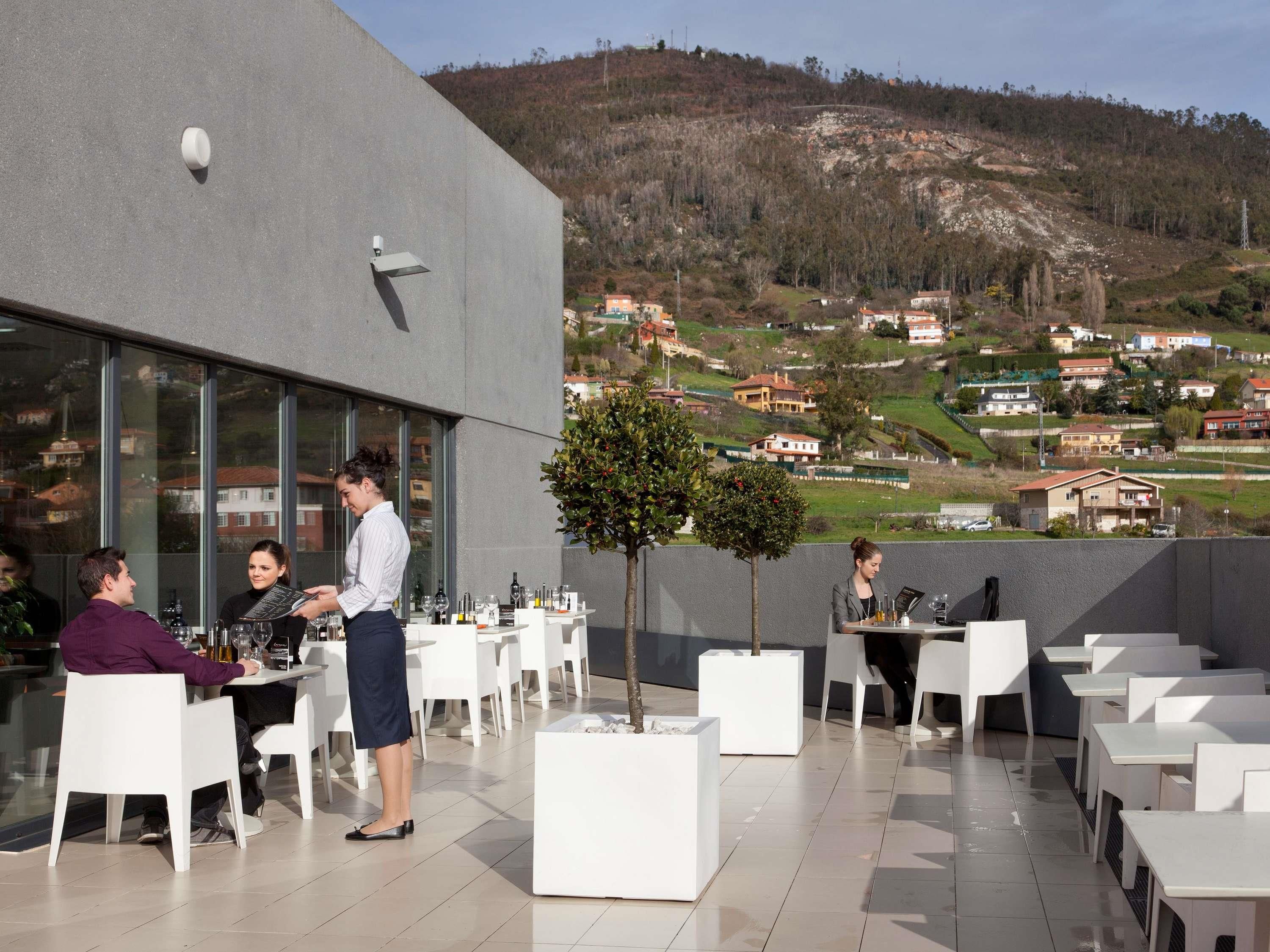 Un área de comedor al aire libre con mesas y sillas, un camarero atendiendo a los huéspedes y vistas a un pueblo en la colina.