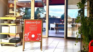 Un área de desayuno con un cartel que indica horario de desayuno temprano, un carro con vajilla y una vista a través de la puerta.
