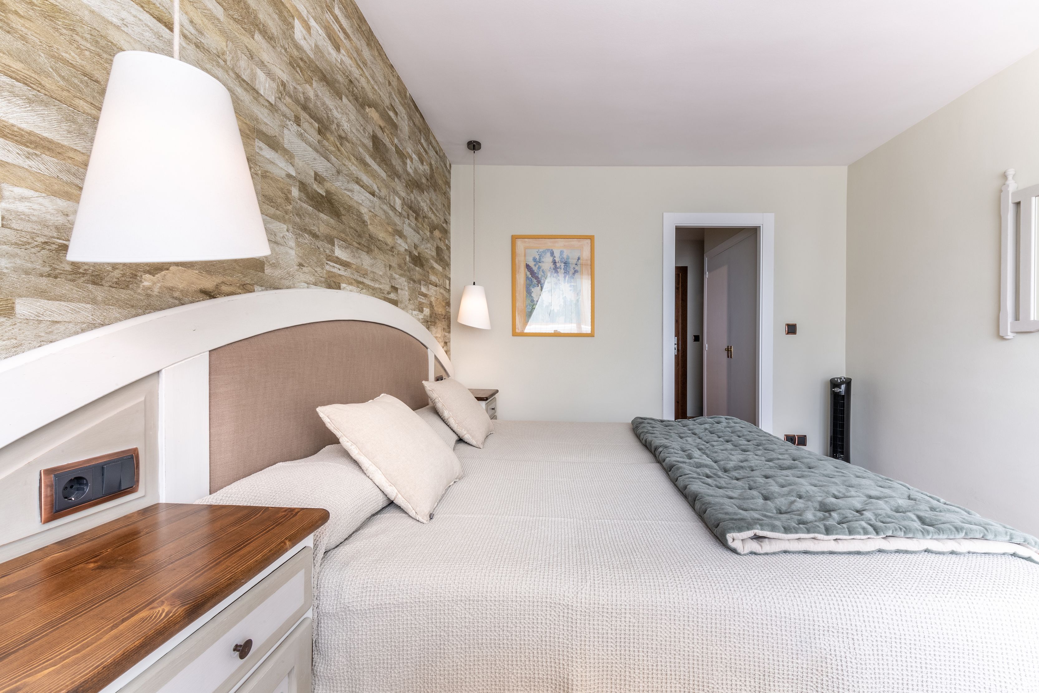 Un dormitorio con cabecera de madera, una mesita de noche, una cama con manta gris, una lámpara colgante y un cuadro en la pared.