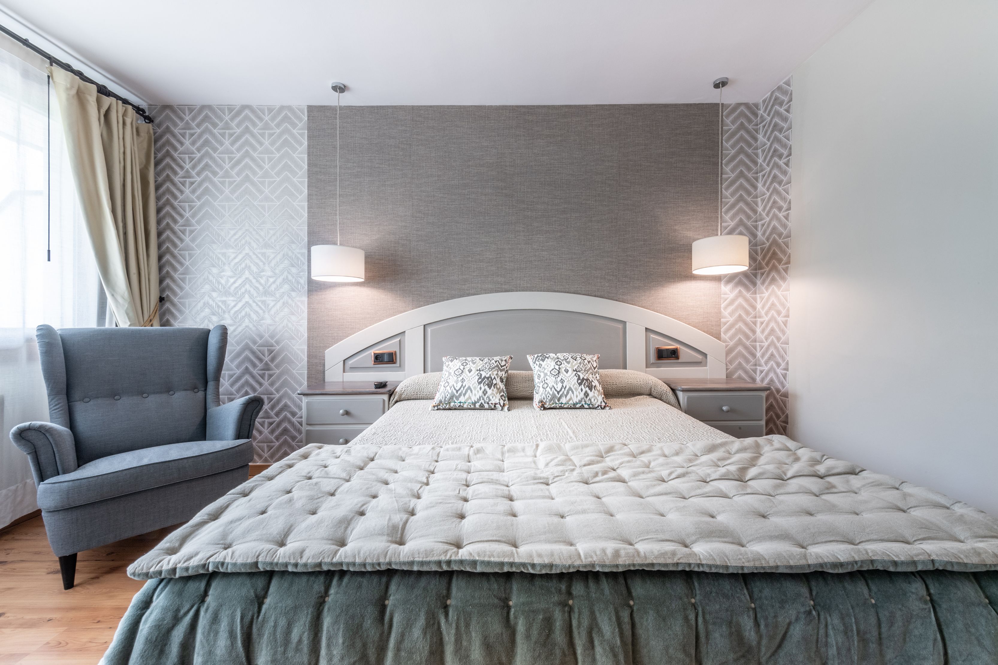 Un dormitorio moderno con una cama capitonada, dos lámparas de noche, un sillón gris y papel tapiz estampado.