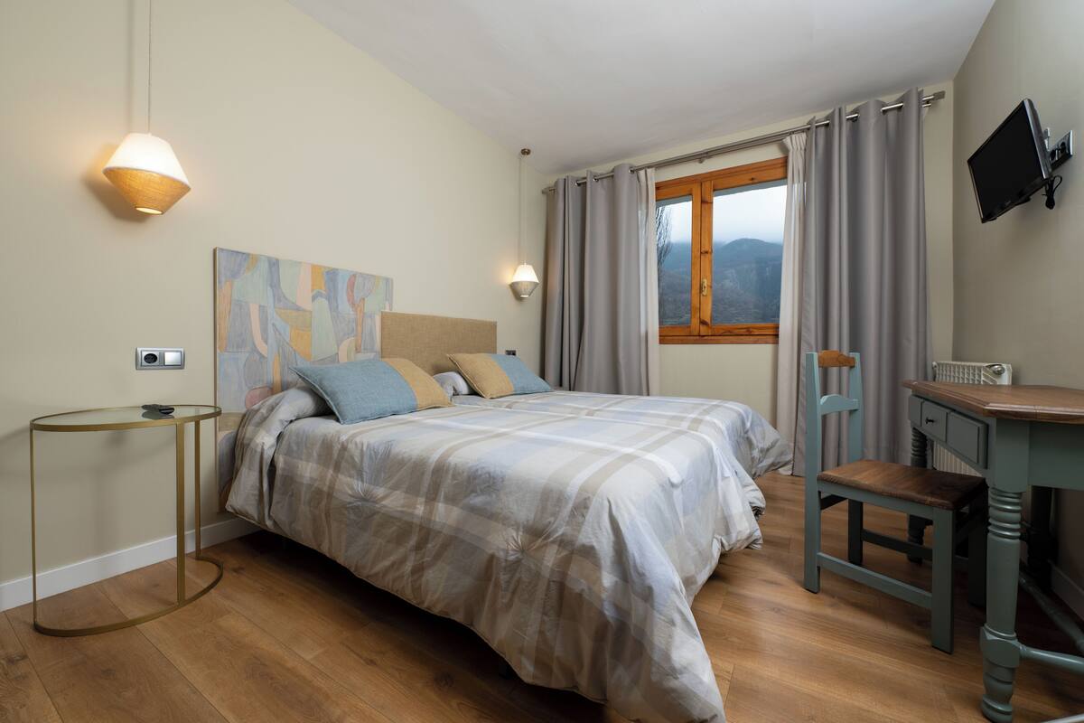 Un dormitorio con una cama, un escritorio, una silla, una lámpara, un cuadro y una ventana con cortinas.