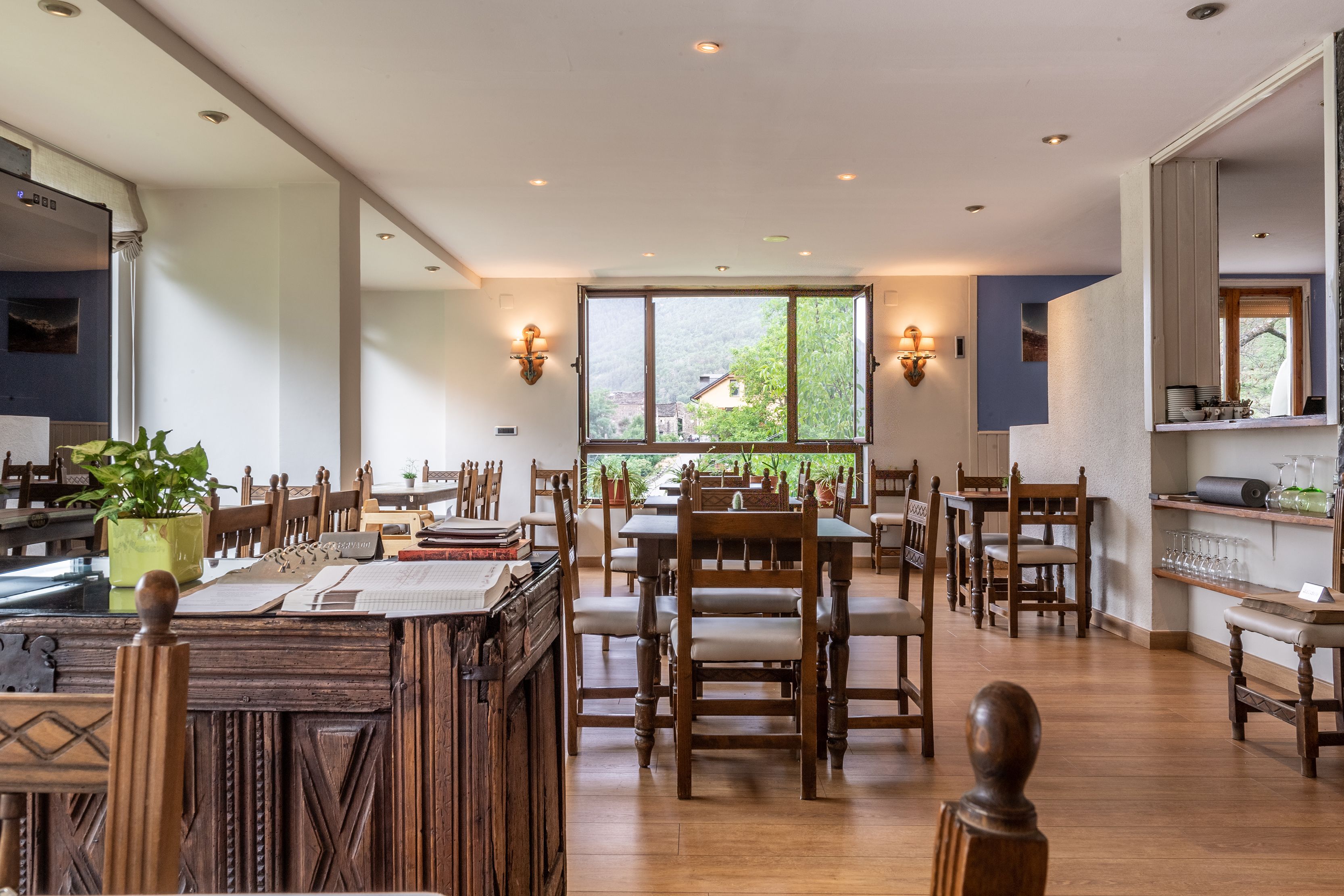 A spacious dining area with wooden tables and chairs, large windows with a view of greenery, and wall-mounted lighting.