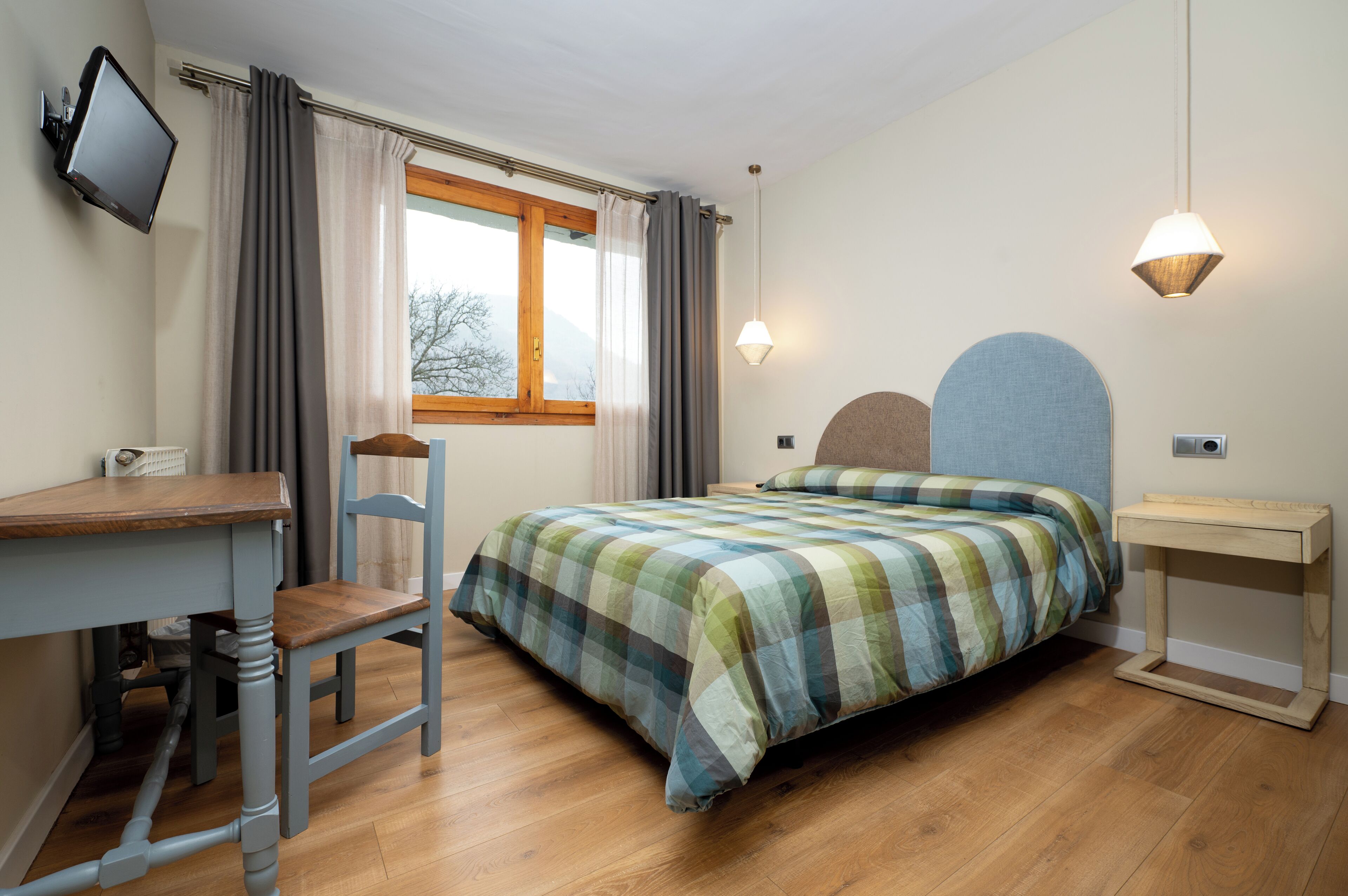 A bedroom with a bed, a desk, a chair, a TV, and a bedside table.