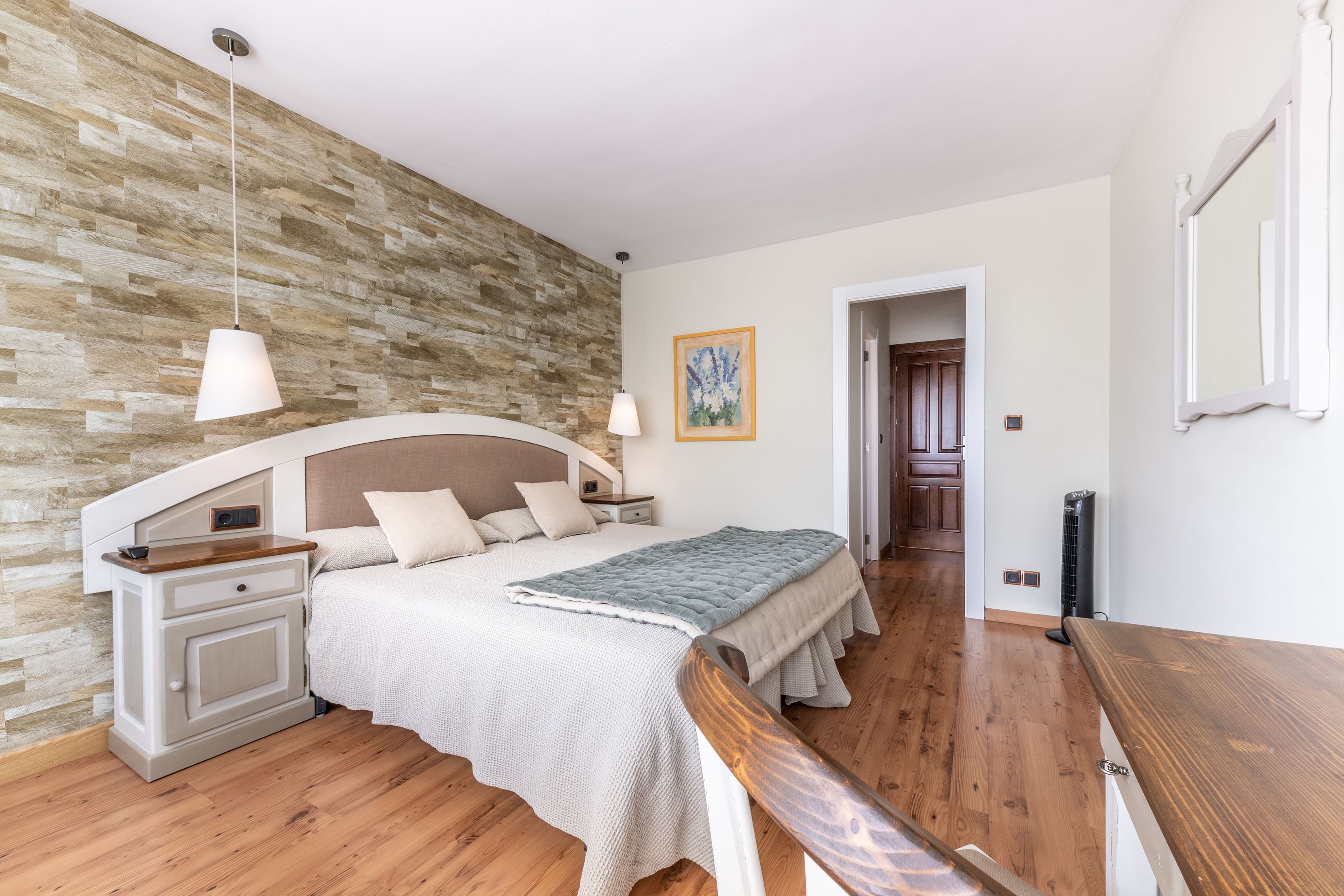 Un dormitorio con cabecera de madera, una cama con edredón blanco, una mesita de noche, un banco de madera, una lámpara de pared y un cuadro enmarcado en la pared.