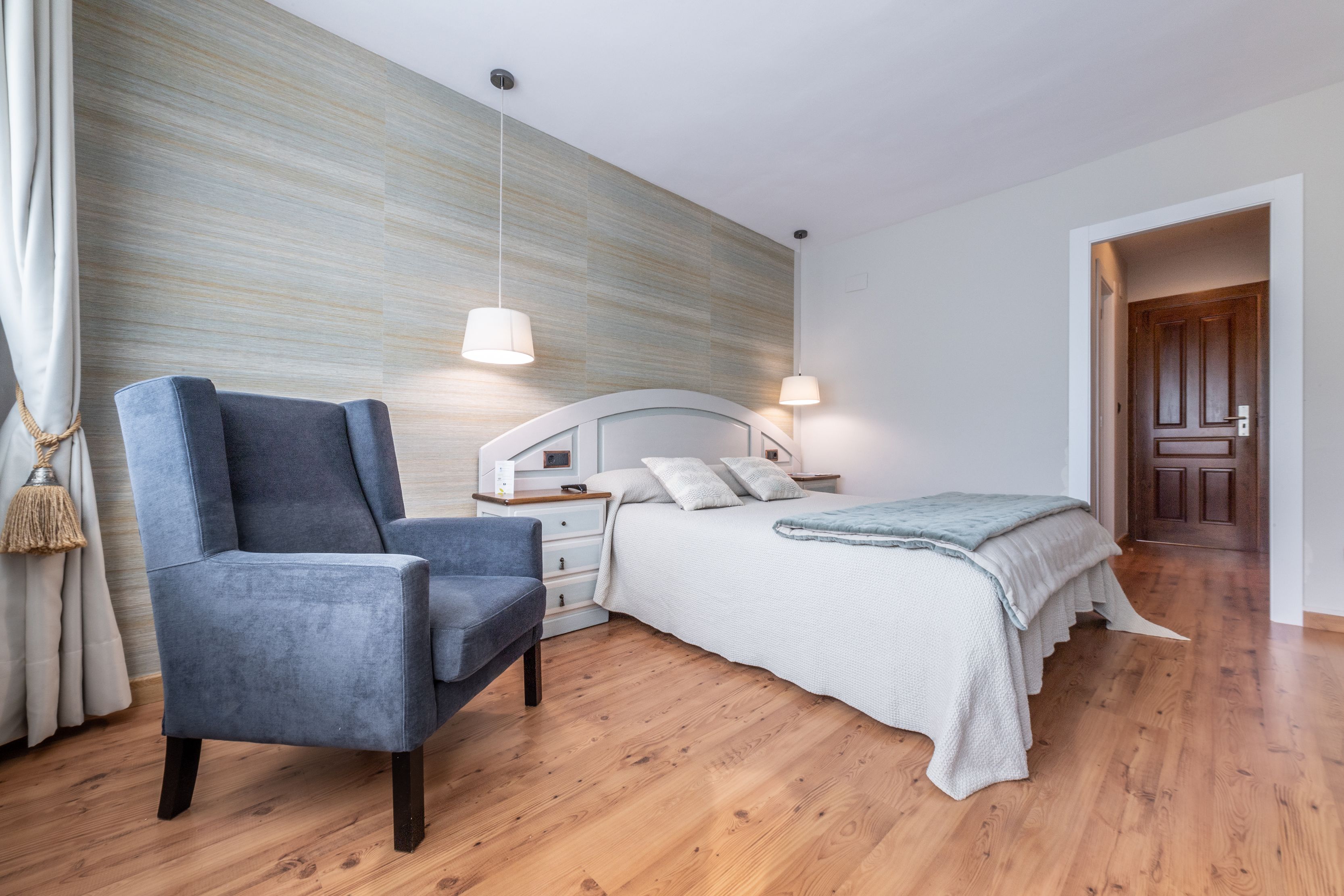 A modern bedroom with a wooden floor, a blue armchair, a white bed with a grey blanket, and a wooden door in the background.