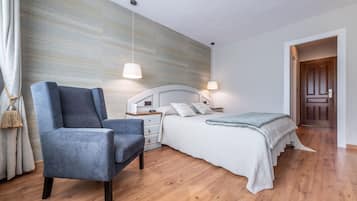 Un dormitorio moderno con suelo de madera, un sillón azul, una cama blanca con manta gris, y una puerta de madera al fondo.