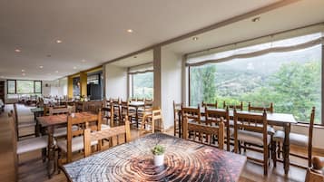 A spacious dining area with wooden tables and chairs, large windows offering a view of a mountainous landscape.