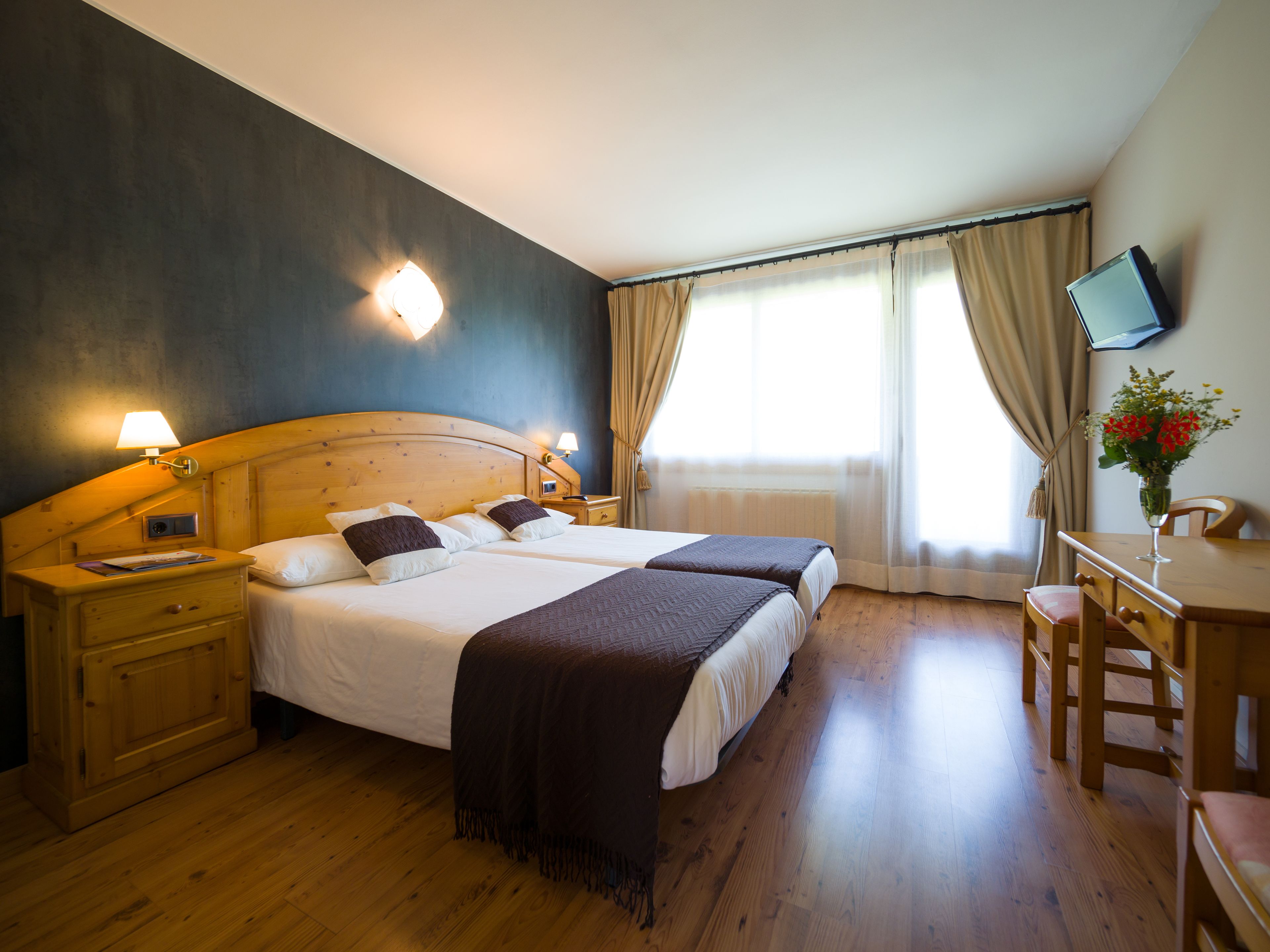 Habitación de hotel con dos camas, cabeceros de madera, lámparas de noche, un escritorio de madera con una silla, un televisor montado en la pared y una ventana con cortinas.