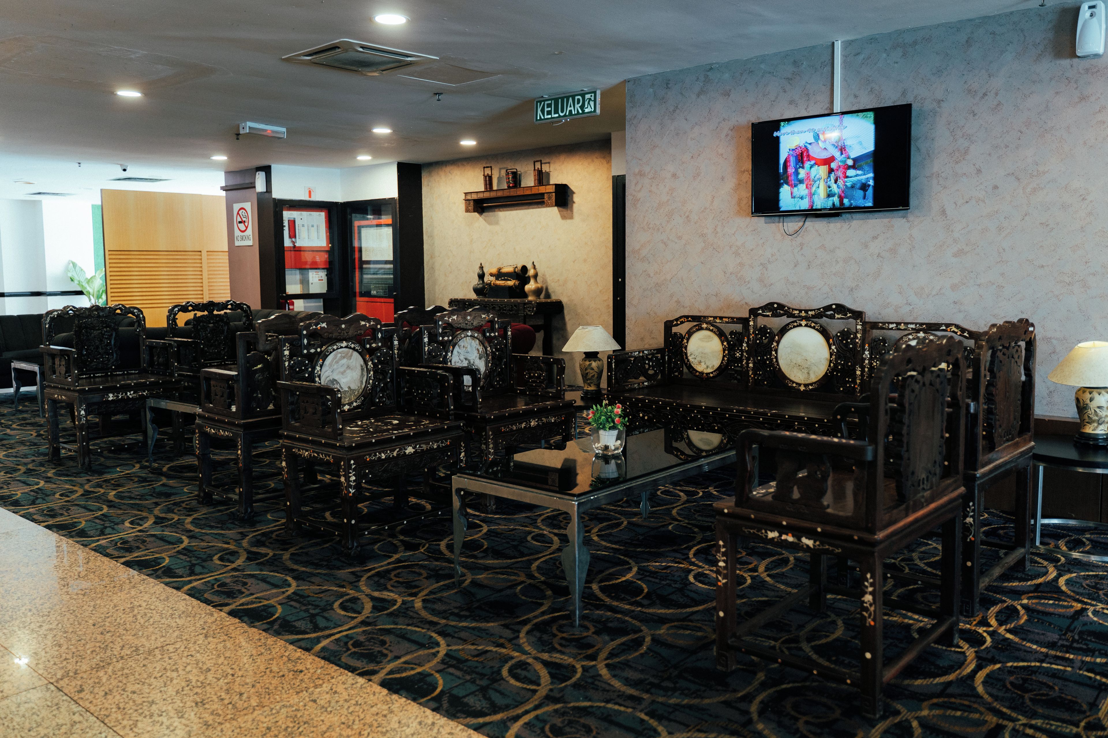 A hotel lobby with ornate wooden furniture, a television mounted on the wall, and a 'KELUAR' sign indicating the exit.