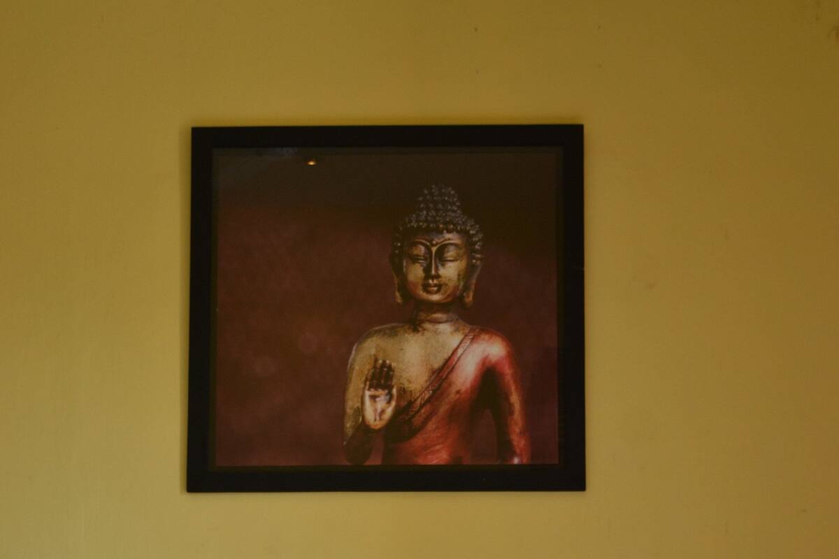 A bed with white bedding and two blue pillows, a framed Buddha statue on the wall, and a yellow wall.