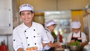 Un chef con uniforme blanco sostiene un plato de comida, mientras otros chefs trabajan en el fondo de una cocina.