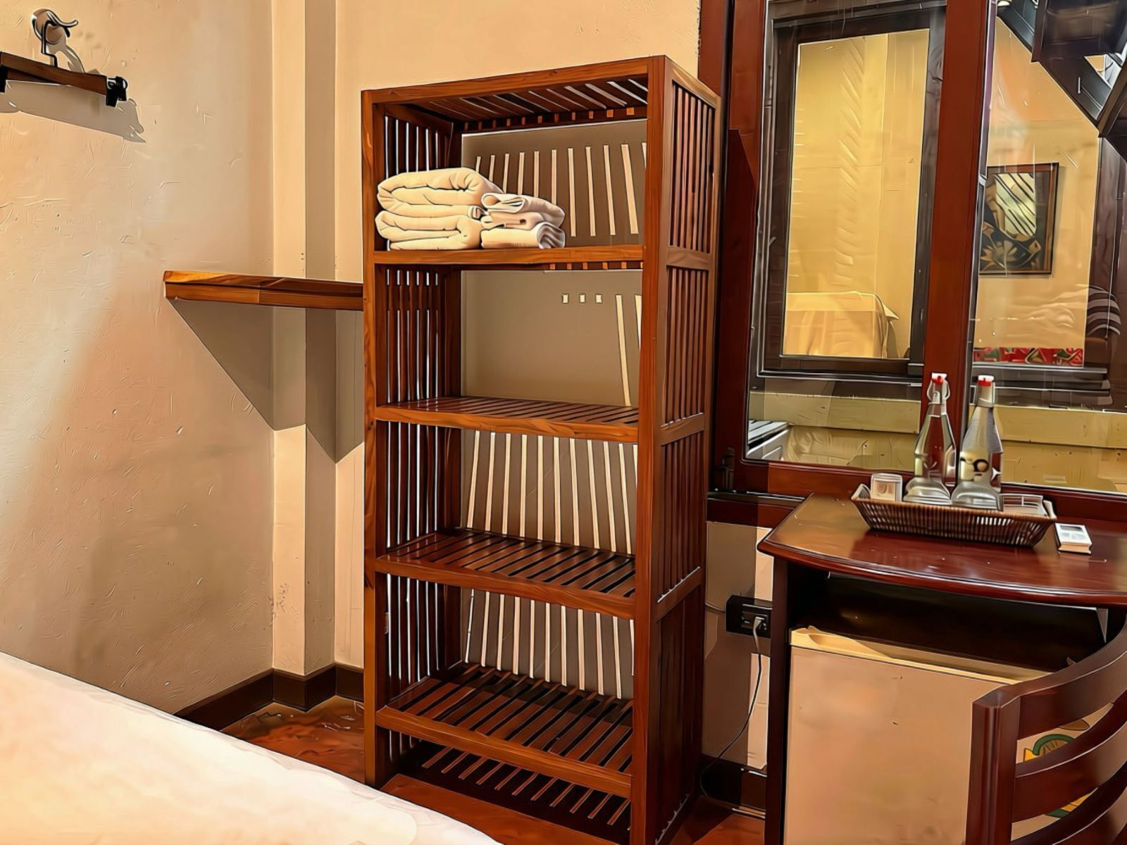 A hotel room with a wooden shelving unit, a small table with a bottle and a book, and a mirror reflecting the room.