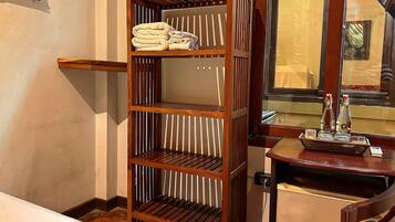 A hotel room with a wooden shelving unit, a small table with a bottle and a book, and a mirror reflecting the room.