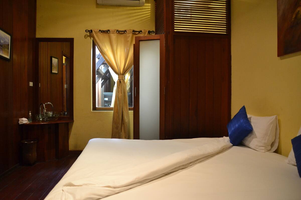 A bedroom with a bed, a window with curtains, a bedside table, and a wall-mounted air conditioner.