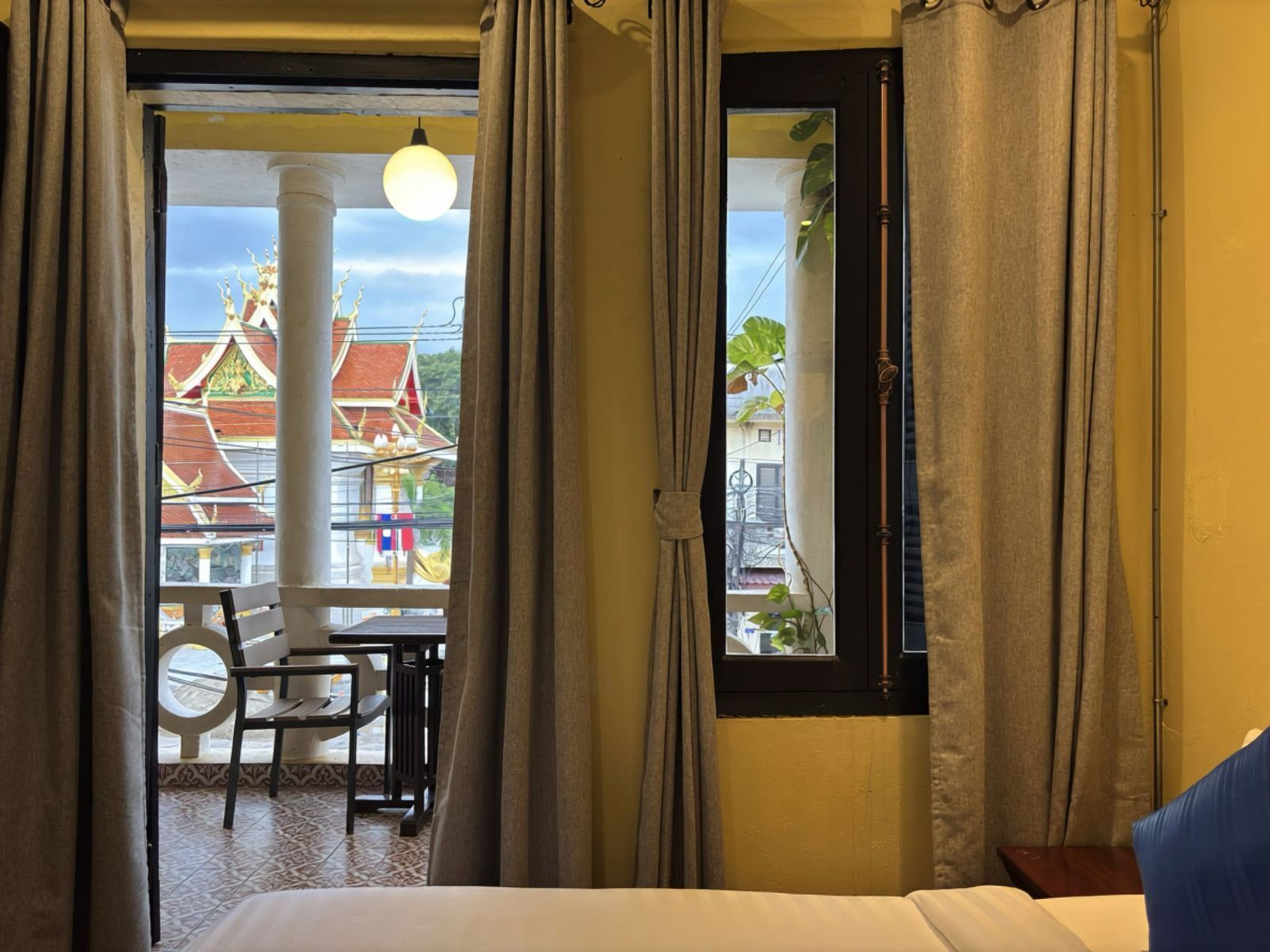 A hotel room with a bed, a window with curtains, a view of a traditional building, and a balcony with a table and chairs.