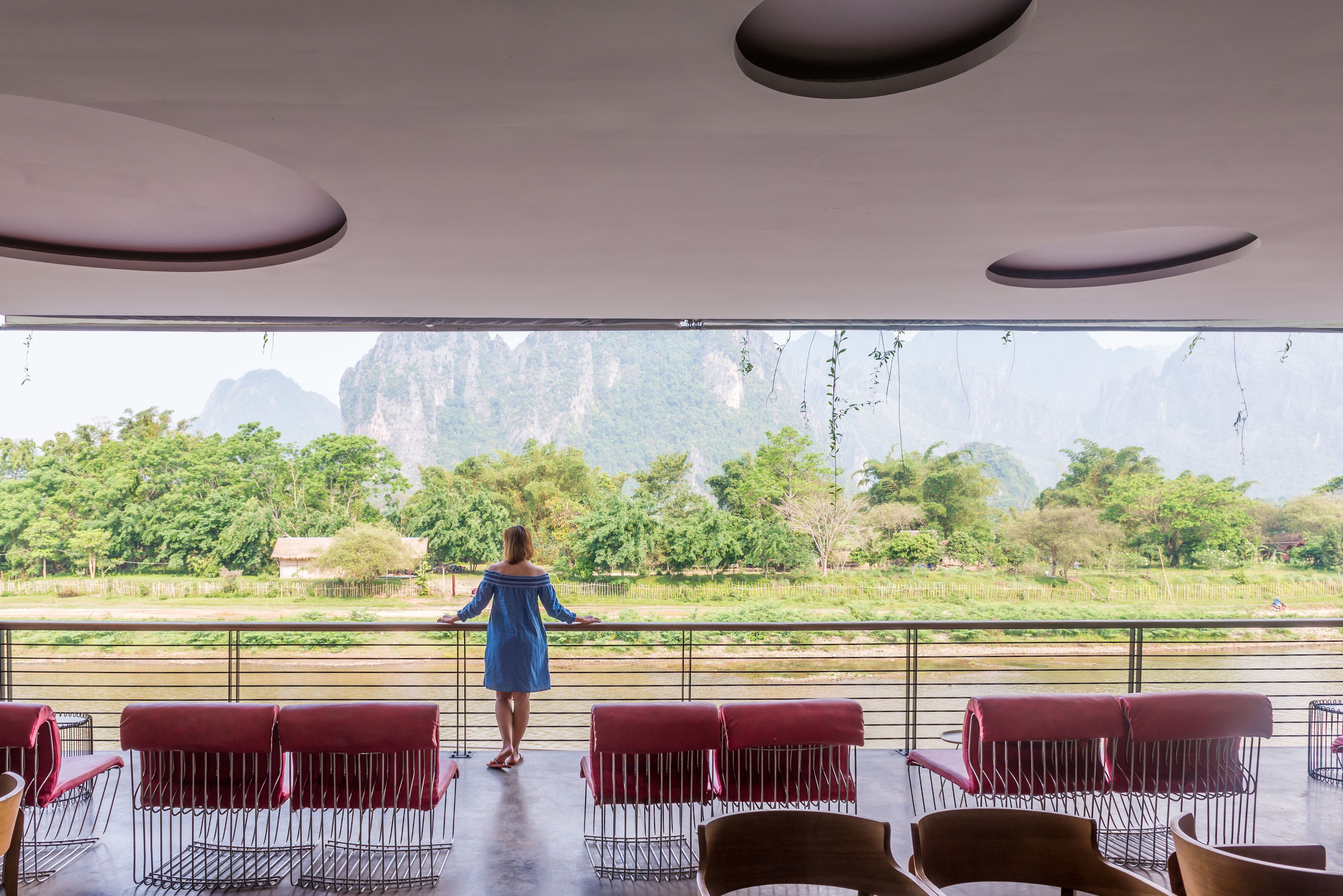 Mulher ao lado de um corrimão com vista para um rio e vegetação, com cadeiras vermelhas e um design moderno no teto.