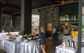 Un comedor con mesas redondas cubiertas con manteles, una pared de piedra y una cocina a la vista al fondo.