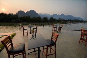 Un área de comedor al aire libre con mesas y sillas, con vista a un río con barcos y montañas al fondo.