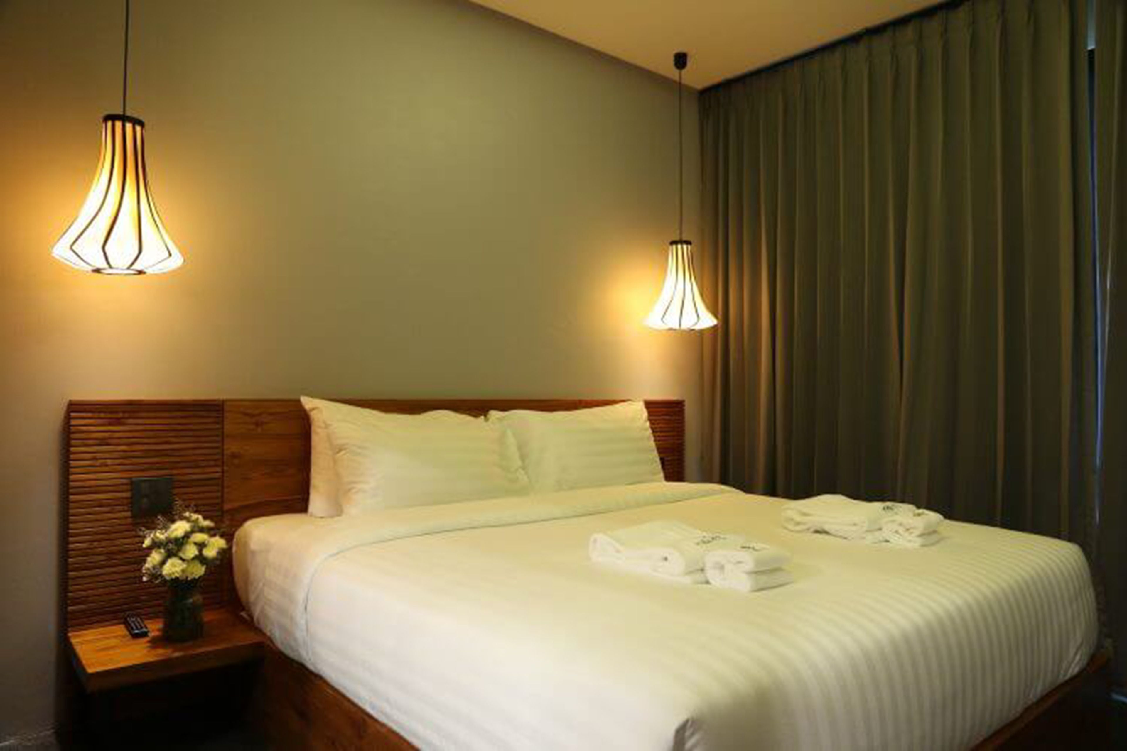 Habitación de hotel con cabecera de madera, cama con ropa de cama blanca, dos lámparas de noche y una mesita de noche con un florero.