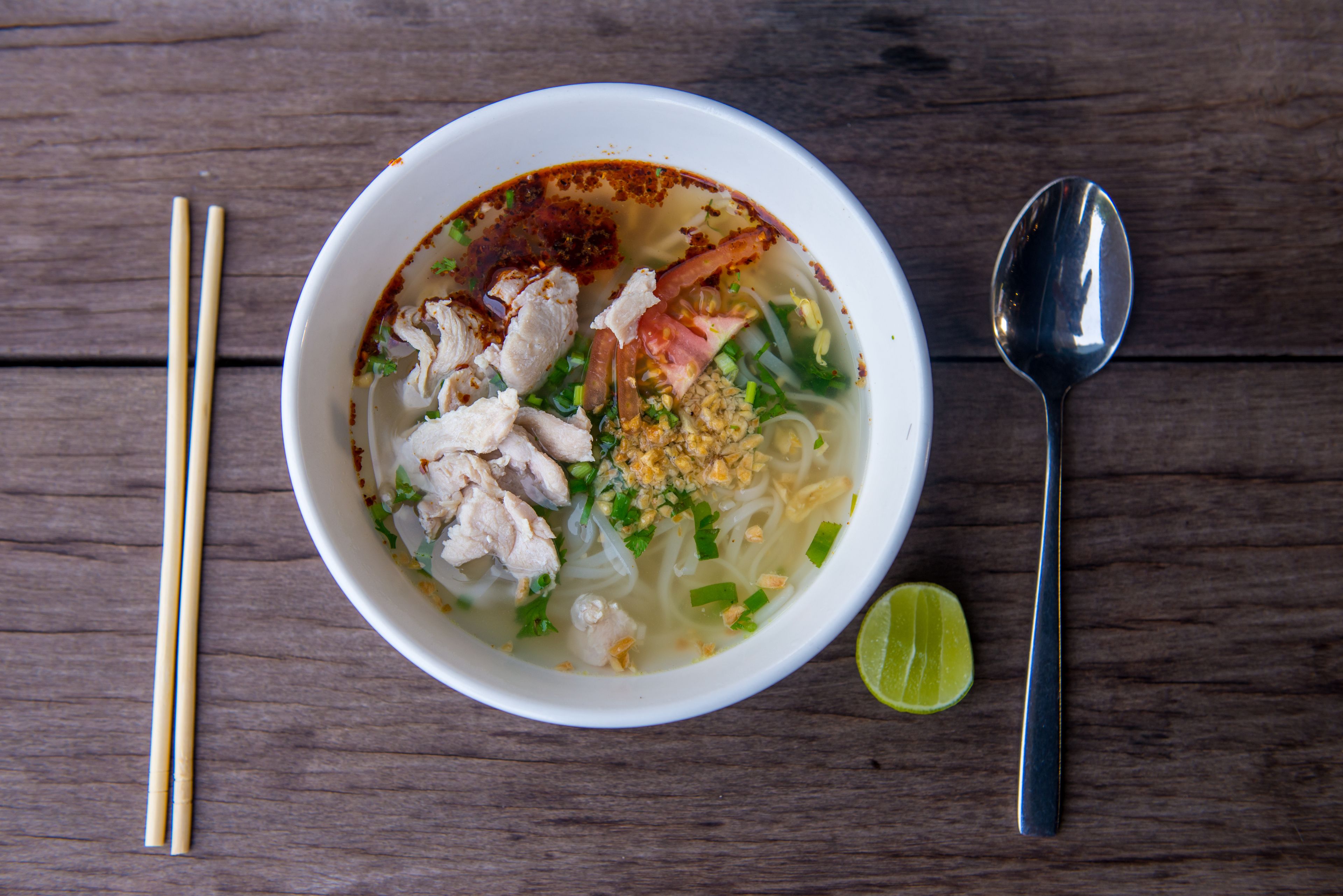 A bowl of noodle soup with various toppings, a spoon, chopsticks, and a lime wedge on a wooden table.
