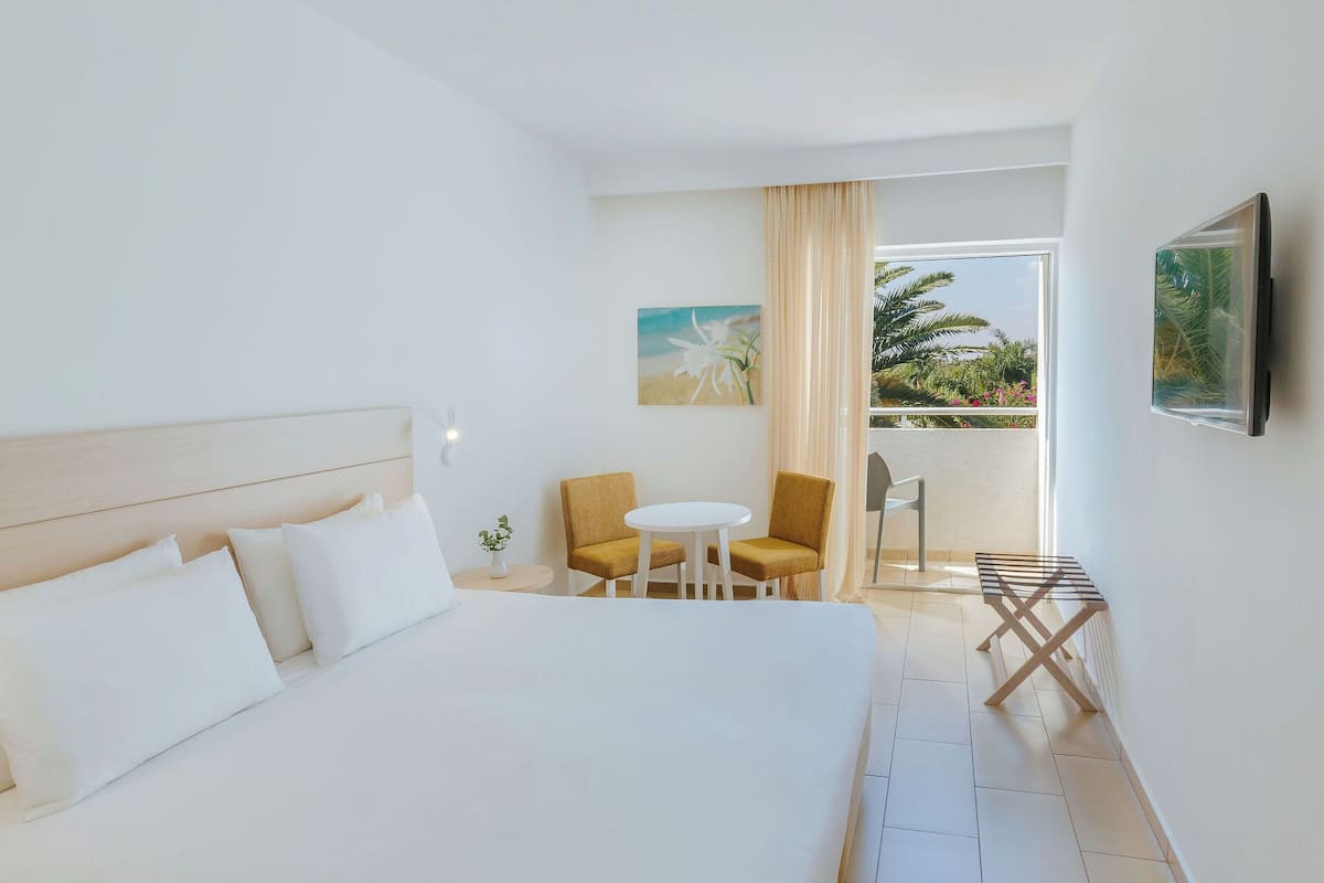 Ein Hotelzimmer mit einem großen Bett, einem kleinen Tisch mit zwei Stühlen, einem an der Wand montierten Fernseher und einem Balkon mit Blick auf Palmen und Blumen.