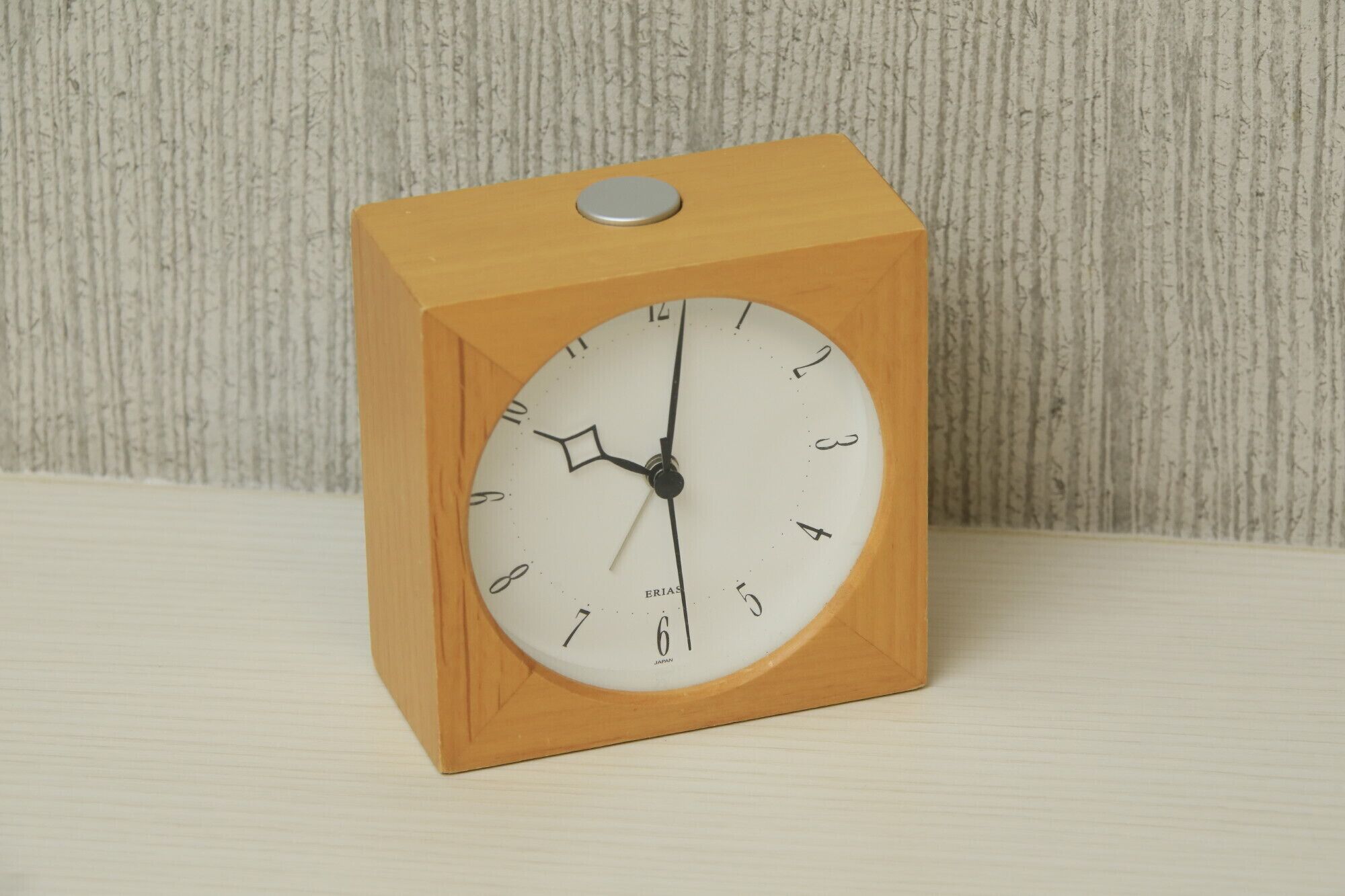 Un reloj despertador de madera con esfera blanca y números negros, sobre una superficie de tonos claros.
