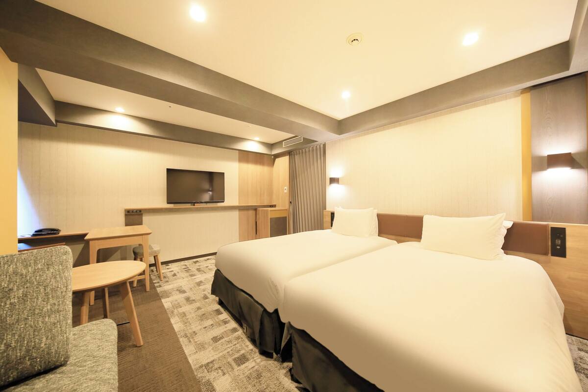 Une chambre d’hôtel avec deux lits, un bureau, une chaise, une télévision et une lampe fixée au mur.