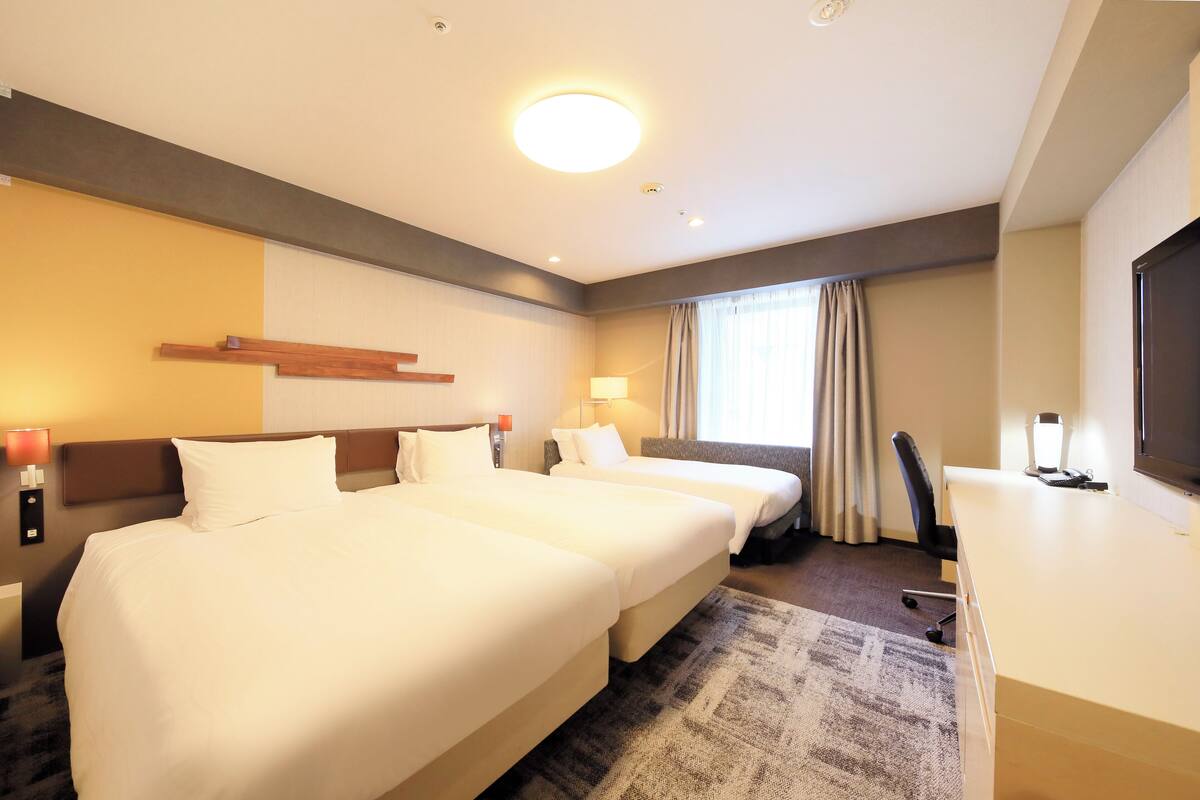 Une chambre d’hôtel avec trois lits, un bureau, une chaise, une télévision et une fenêtre avec des rideaux.