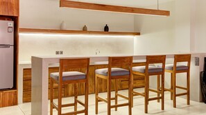 A modern kitchen with wooden bar stools, a built-in refrigerator, and a shelf with decorative items. - One Eleven Haven (Keroboken Kelod)
